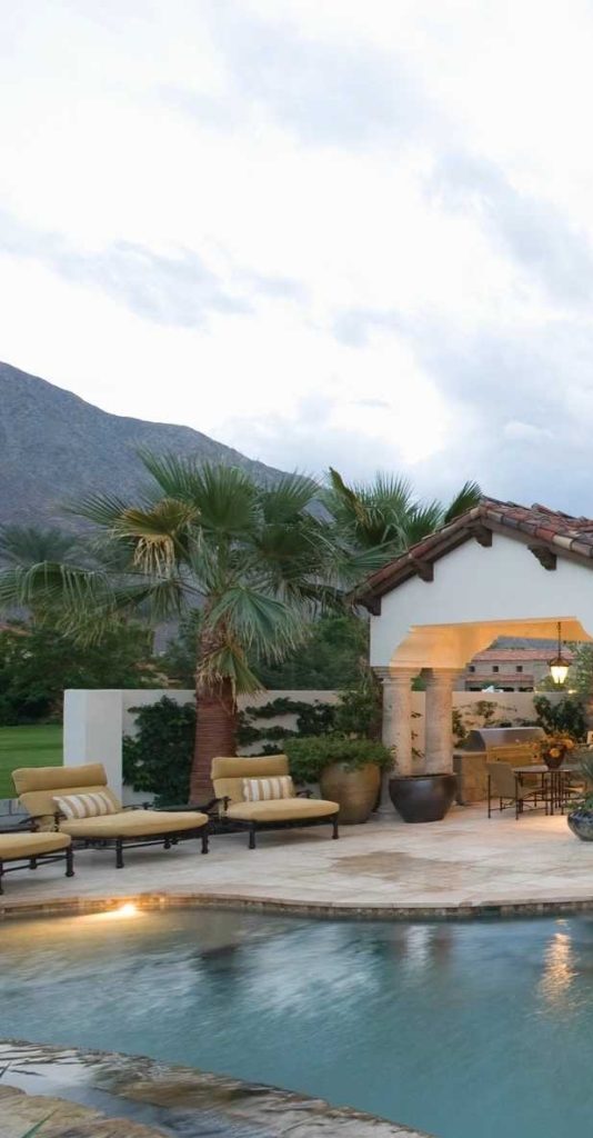 A luxurious outdoor patio with lounge chairs by a pool, palm trees, and a covered seating area. Mountains and a cloudy sky are visible in the background.