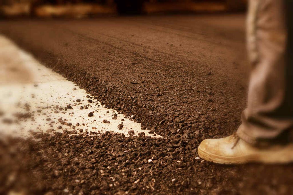 Close-up of a person’s boot standing on freshly laid asphalt, next to a white edge line, with rough black gravel covering the ground. The upper body of the person is out of frame.