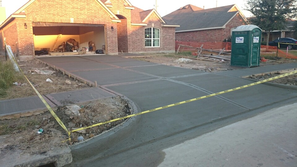 Freshly poured concrete driveway in front of a brick house under construction, with caution tape around the area and a portable toilet nearby. The garage is open, showing construction materials inside.