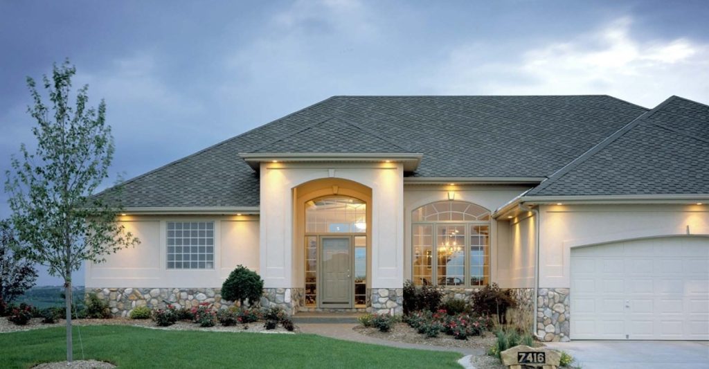 Single-story house with large windows, stone accents, and a neatly landscaped front yard. Outdoor lights are on, and the sky is overcast. The garage door is on the right, with a small tree and garden visible in front.