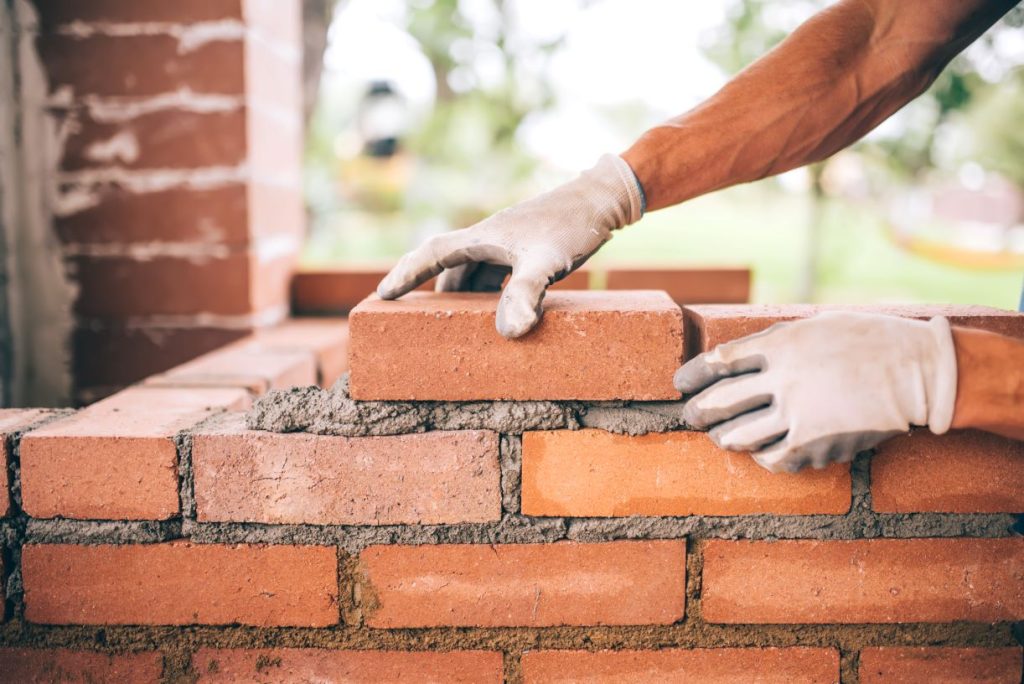 A person wearing white gloves is laying red bricks with cement, building a brick wall outdoors. The scene is close-up, showing hands carefully placing a brick in line with others.