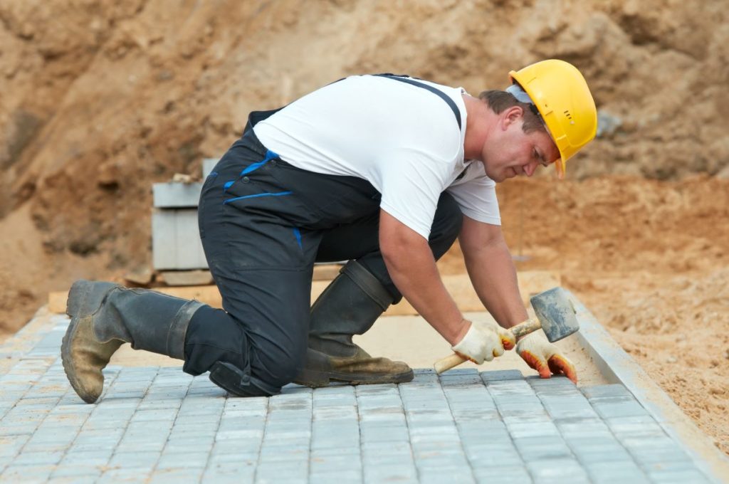 A construction worker wearing a yellow hard hat, gloves, and boots is kneeling on the ground, using a rubber mallet to lay paving stones on a construction site with a dirt background.