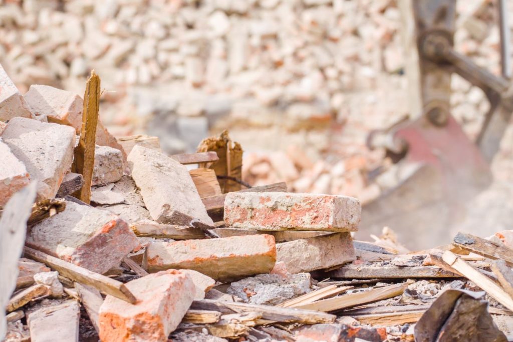A pile of broken red and white bricks mixed with wooden debris, with a blurred background of more rubble and a construction machine arm partially visible on the right.