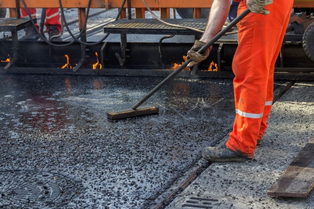 A worker in orange safety clothes smooths hot asphalt on a road using a tool, with flames visible from machinery in the background.