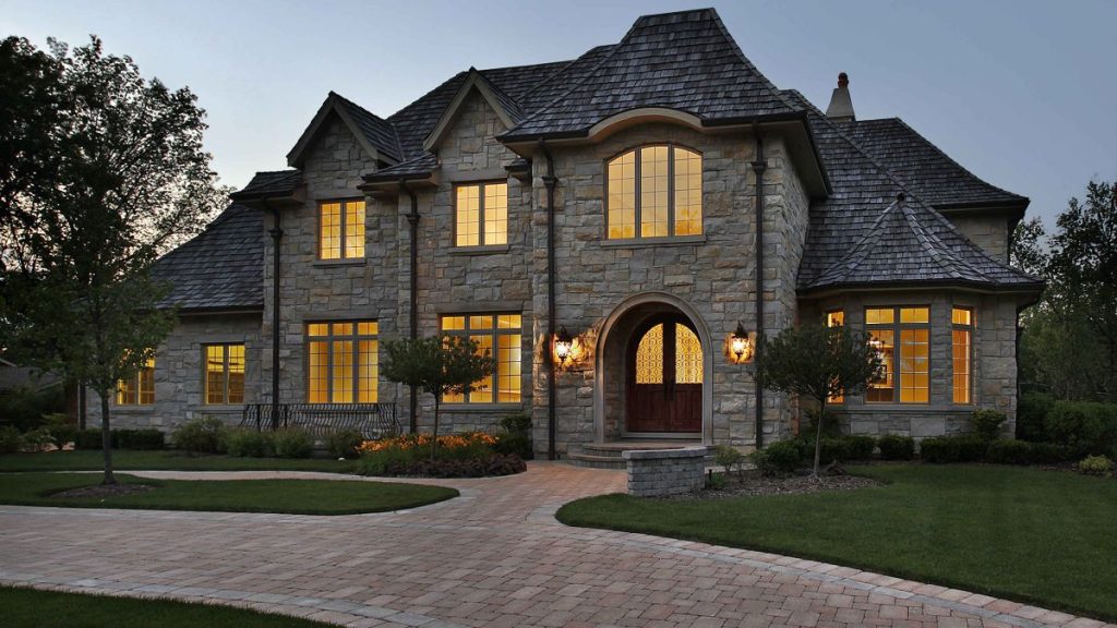 A large stone house with multiple gables and lit windows at dusk, featuring a wooden front door, arched entryway, lantern-style lights, manicured lawn, trees, and a curved brick driveway.