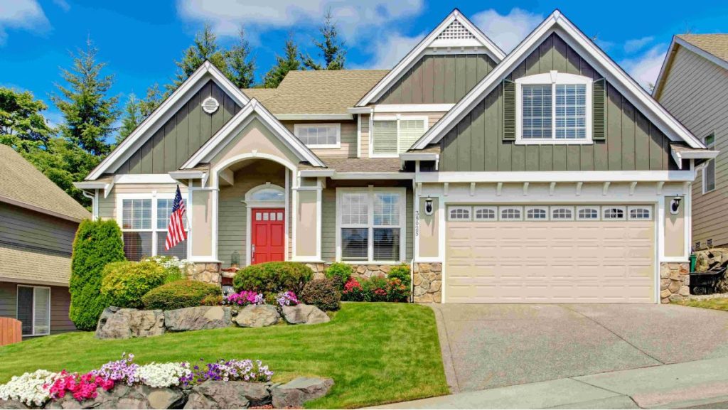 A two-story suburban house with gray siding, white trim, a red front door, and a two-car garage. The yard is landscaped with green grass, flower beds, rocks, and an American flag by the entrance.