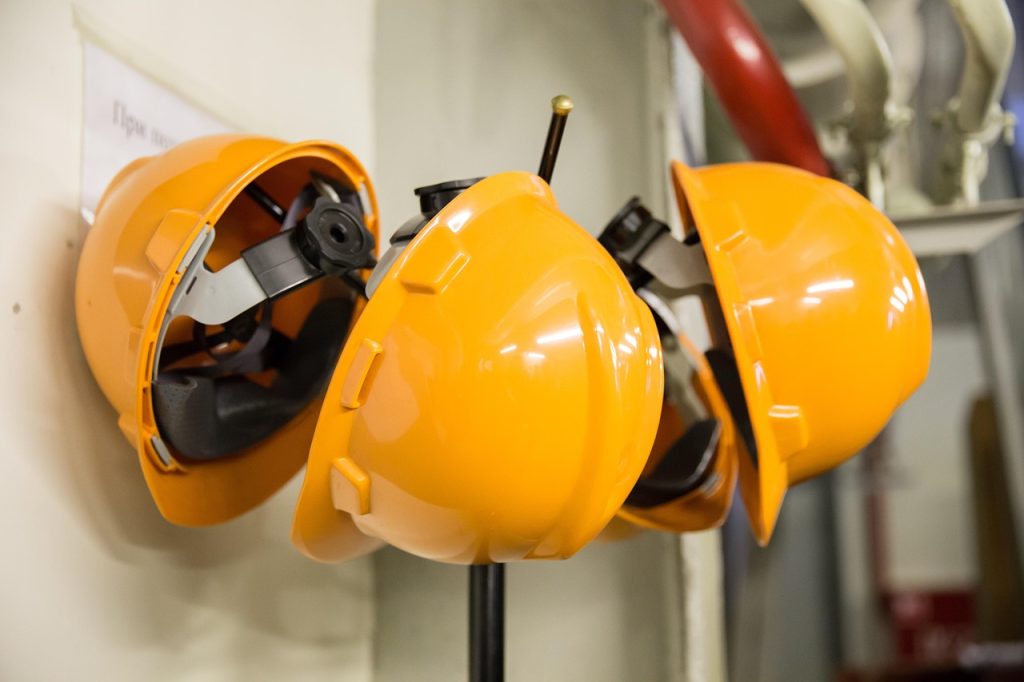 Three bright orange safety helmets are hanging on hooks against a light-colored wall, with part of some industrial pipes visible in the background.