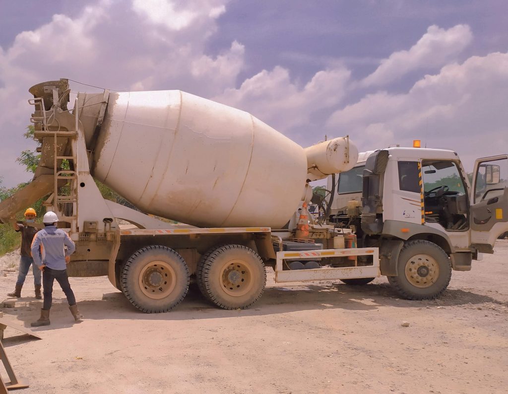 A cement mixer truck is parked on a construction site with three workers in safety gear standing nearby under a partly cloudy sky.
