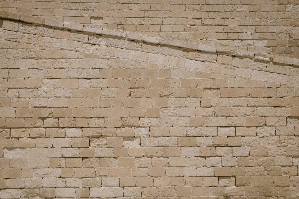 A beige stone brick wall with a diagonal line of lighter-colored bricks running across it from the upper left to the lower right. The wall has a rough, textured surface and irregular brick shapes.