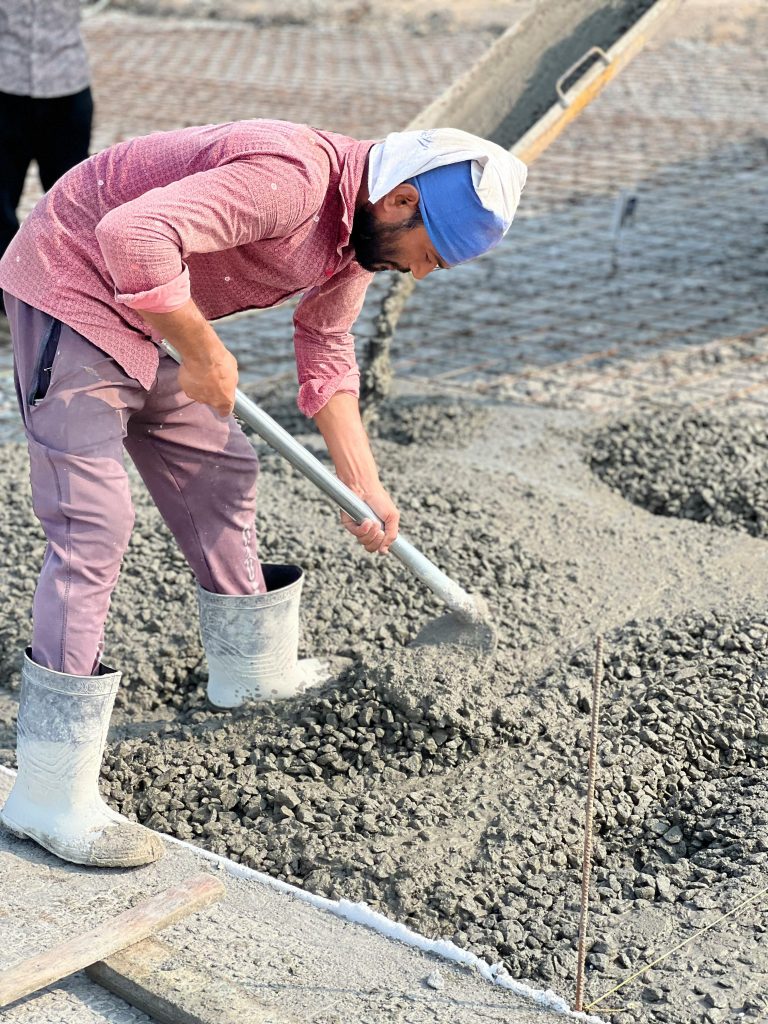 A construction worker in a pink shirt, lavender pants, white boots, and a blue head covering spreads wet concrete with a shovel on a building site.
