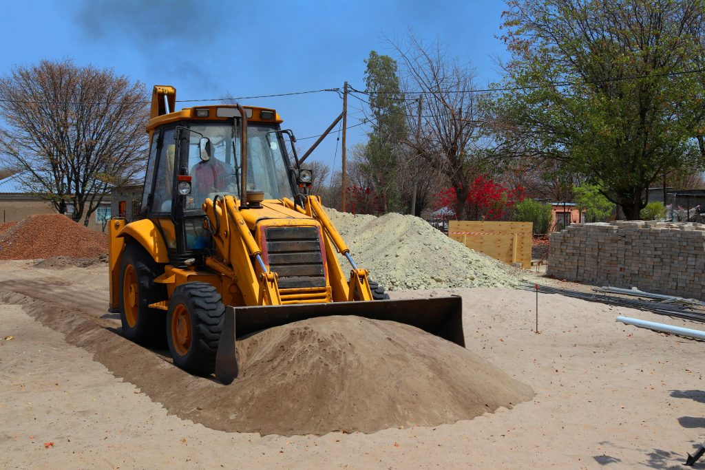 A yellow backhoe loader is parked on a construction site, with its front bucket filled with sand. Piles of materials, trees, and construction supplies are visible in the background.