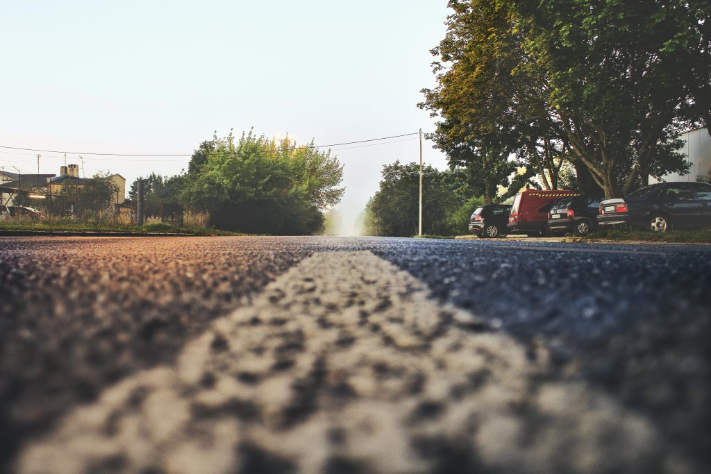 A low-angle view of a paved road with a white line in the center, leading toward trees and parked cars on the right side. The scene is calm with soft, natural lighting.