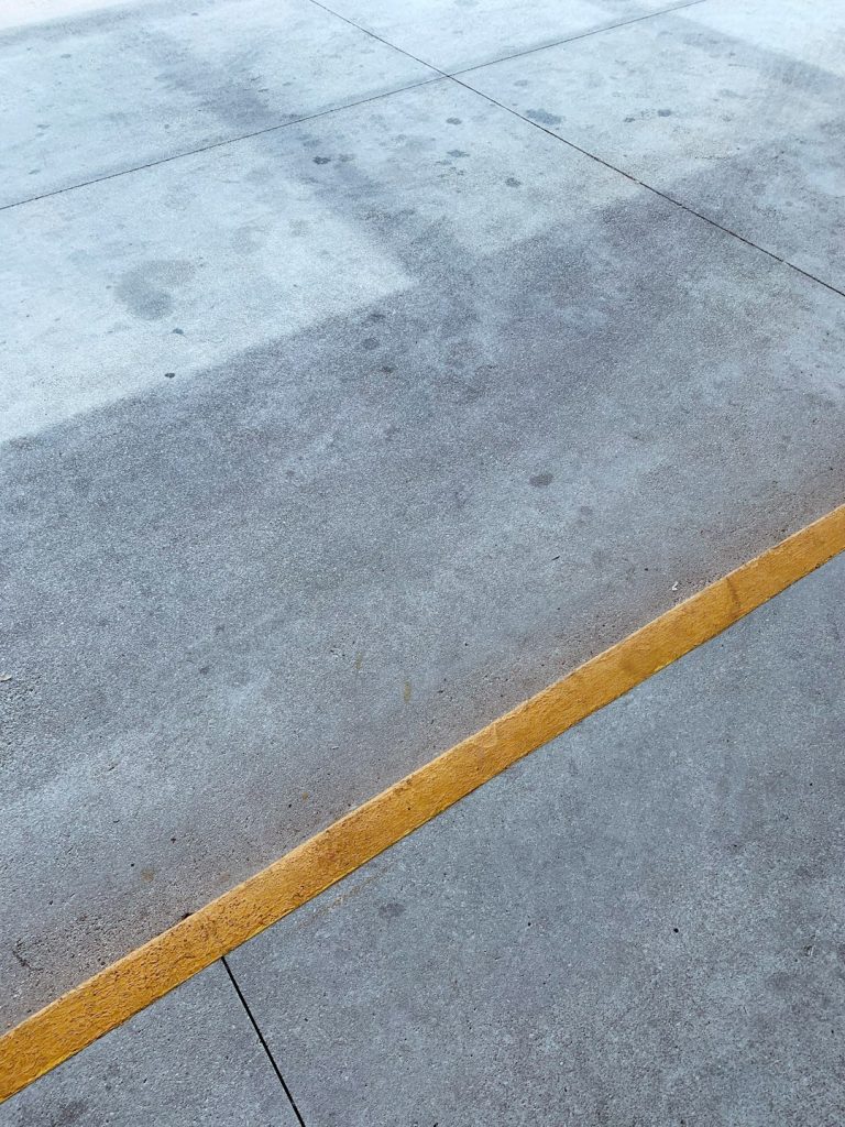 A concrete pavement with a single yellow line running diagonally across it. The surface shows some faint stains and marks, and is divided by straight, narrow grooves.