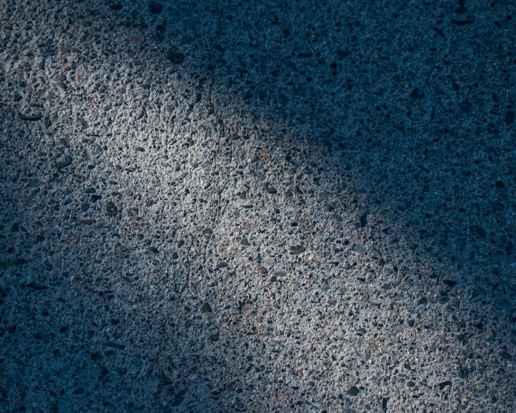 A close-up of a textured concrete surface with scattered small pebbles, partially covered by a shadow, creating a contrast between light and dark areas.
