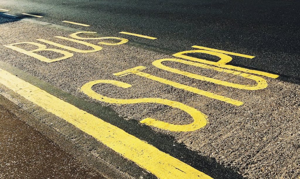 Yellow BUS STOP letters painted on the asphalt beside a road, with a yellow line marking the edge of the pavement. The image is angled slightly from above.