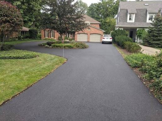 A freshly paved black asphalt driveway curves between green lawns and landscaping, leading to a brick house with a double garage and a white car parked in front. Another house with a porch is visible to the right.