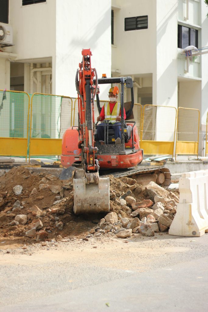 A construction worker operating an orange excavator moves dirt and rocks at a construction site in front of a fenced-off building.