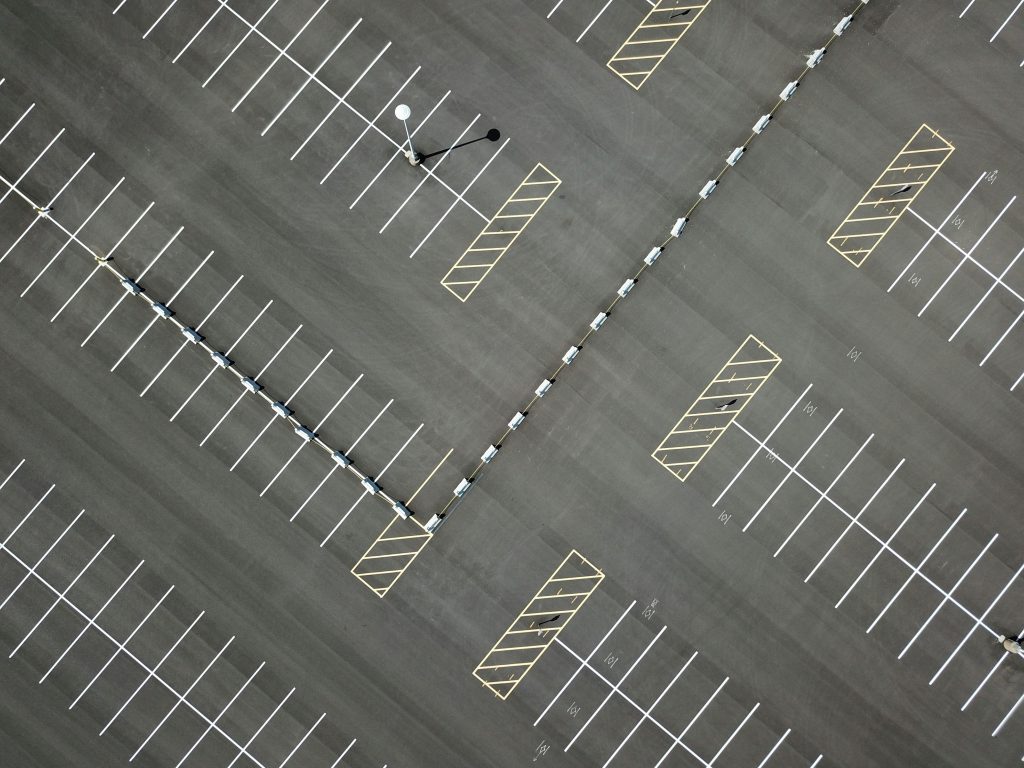 Aerial view of an empty parking lot with white-lined parking spaces, diagonal yellow-striped no-parking zones, a streetlamp, and a dividing barrier running through the center.