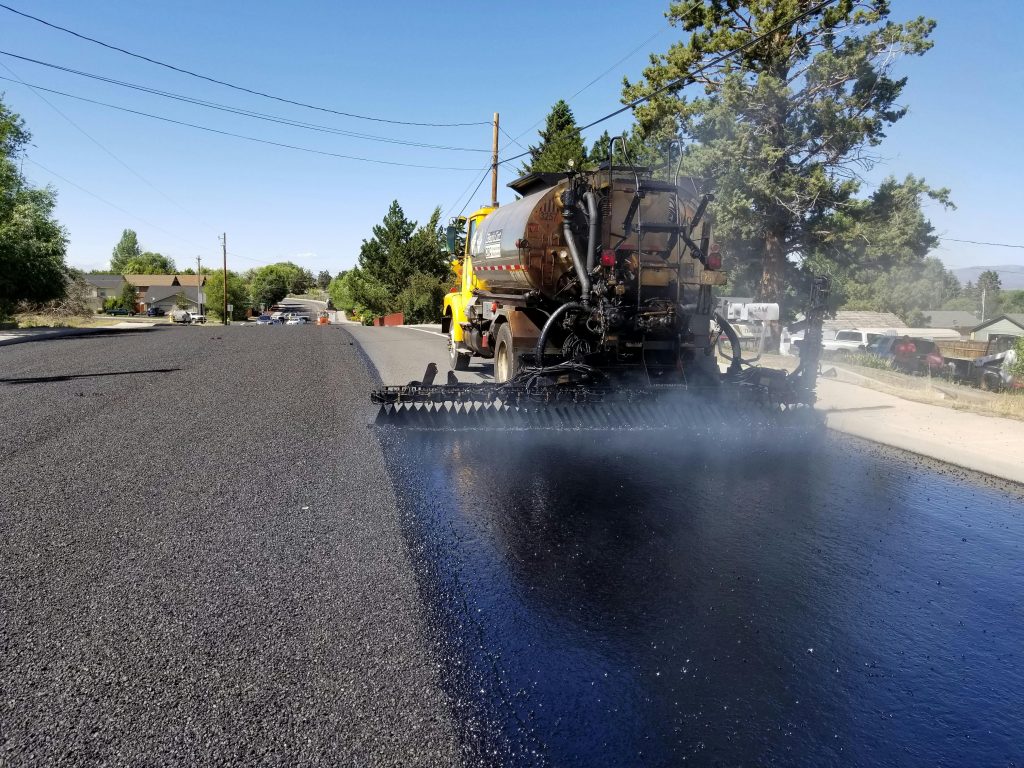 A road construction vehicle sprays liquid asphalt onto a roadway, preparing the surface for paving. Fresh black asphalt covers part of the road, with houses and trees visible in the background under a clear sky.